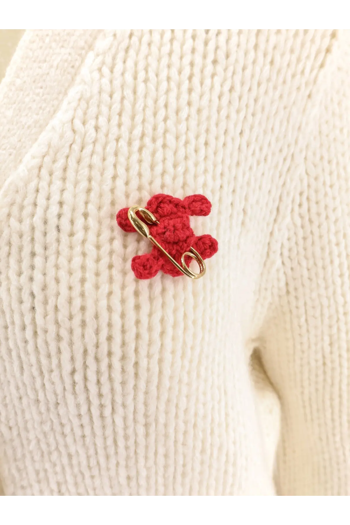 A red crocheted teddy bear pin with a gold safety pin clasp displayed with a celine women’s white silk and cashmere cardigan
