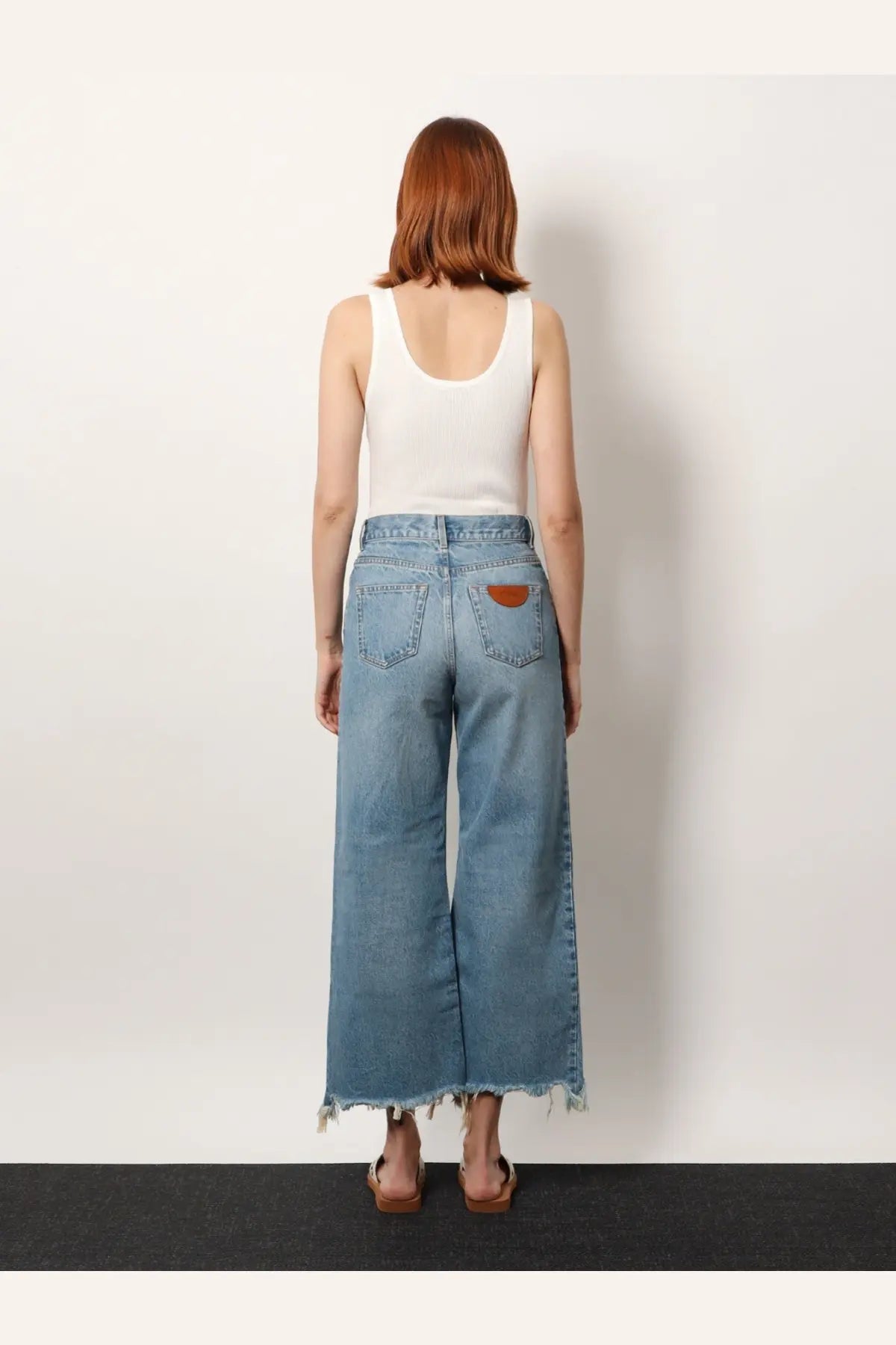 Light-wash denim wide-leg trousers with frayed hem and brown leather patch on back pocket shown from back view