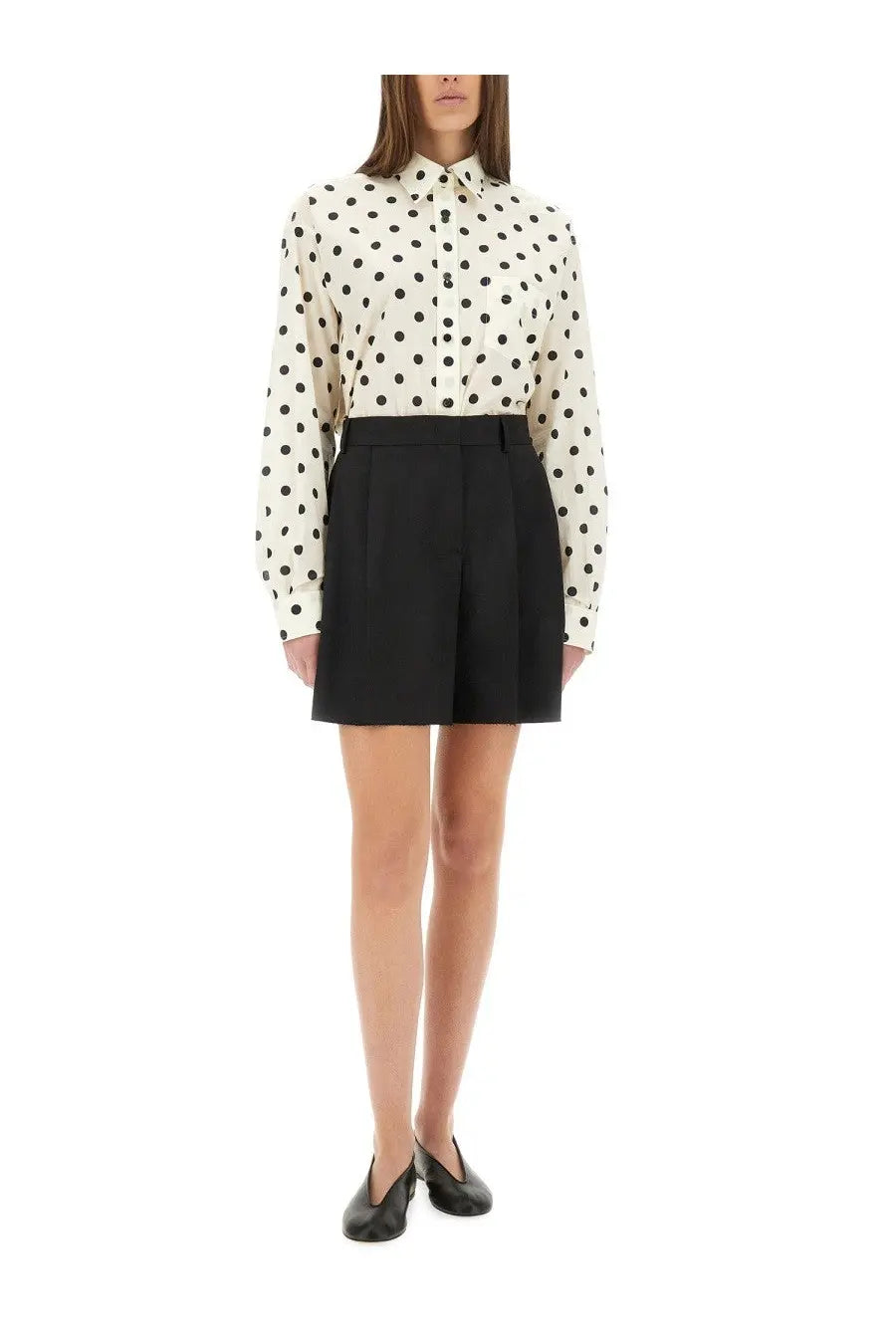 A woman wearing Moschino women’s asymmetrical black wool shorts paired with a cream blouse with black polka dots