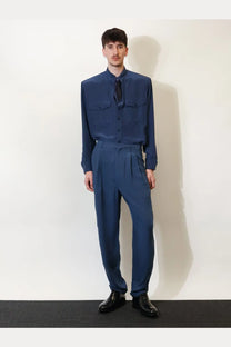 A man wearing Saint Laurent men’s blue silk pleated trousers and a navy blue long-sleeved button-up shirt with chest pockets