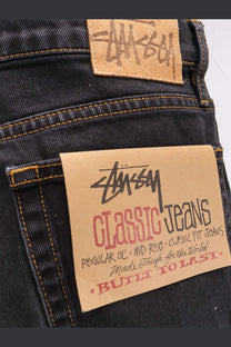Stussy men’s classic dark-wash denim jeans with tan leather patch and matching paper label in black script