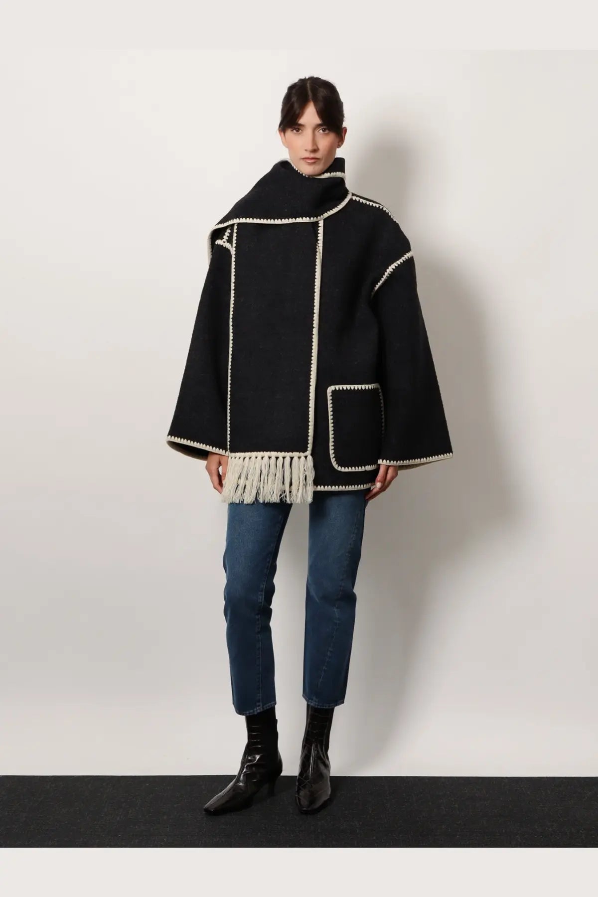 Black woolen cape with cream embroidered trim and fringe detailing shown with Toteme women’s twisted seam jeans blue